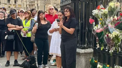 A crowd of people are standing listening to a woman dressed all in black holding a microphone. There is a Labrador on a lead lying on the ground, and there are floral tributes on railings to the right of the screen.
