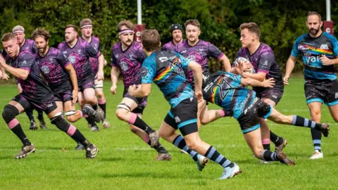 Bristol Bisons RFC Ruby players play on a pitch, with one team member in a blue and rainbow kit tackling another player in a pink and black kit. People are running around them during the match. 