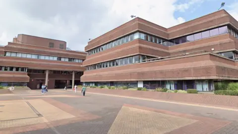 A three-storey modern building with brutalist features. There is a brick-paved open space in front of the building, with people walking across it.