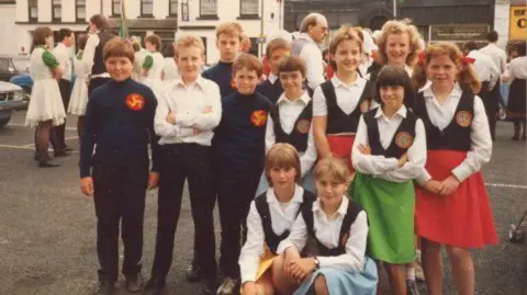 JUAN GARRETT A group of young Manx dancers in costumes. Most are wearing white shits and black waistcoats, wit the girls wearing coloured skirts and he boys black trousers. Three of the boys are wearing dark blue tops with a three legs of Man logo on the breast.