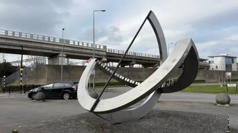 Martin Heath/BBC The approach to a roundabout which is being navigated by a black car. There is a sculpture consisting of two metal semicircles hinged together and a central vertical. It is on the pavement alongside the roundabout. A concrete section of overhead railway line is visible in the background, and a road going under the line can be seen to the left.