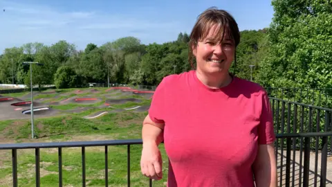A woman in a pink T-shirt with dark hair leans on a railing in front of a bicycle pump track