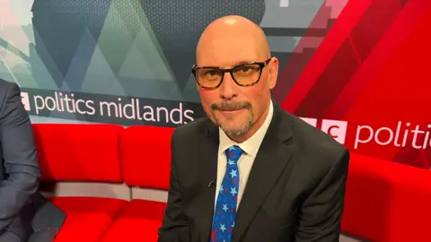 A man sat on a red sofa, wearing a black suit, white shirt and blue tie. 