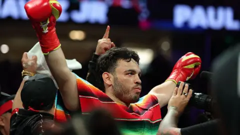 Chavez Jr with his gloved fists in the air as he prepares to fight. He wears a colorful Mexican poncho in the ring
