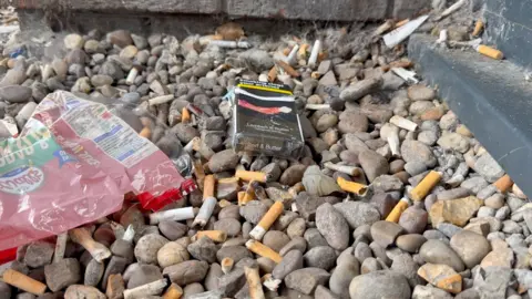 Cigarette butts and empty cigarette packets are discarded on a stony ground outside a hospital.