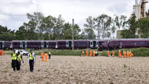 EPA/Shutterstock Two purple, crashed trains in the background, with police and railway workers in orange standing in the field in front of the tracks.