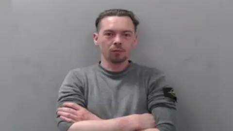 PSNI Aaron Thomas Curragh is wearing a grey jumper and has his arms crossed. He has brown hair. The background is blurred grey