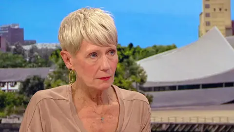 Julie Francis sits looking at the interviewer, who is out of shot, on her right. She has short blonde hair and wears a beige top and gold loopy earrings. The BBC Breakfast background shows the rooftops of various buildings amid the trees.