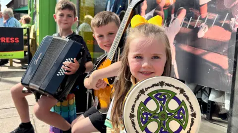 Two boys and a girl are holding instruments and smiling. One has an accordion, one has a guitar and one has a drum. There is a Celtic design on the drum. 