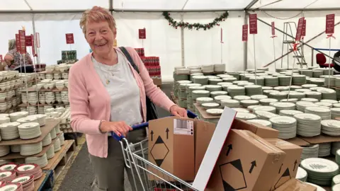 A woman pushing a trolley containing boxes is smiling widely. Behind her are many plates and dishes stacked in a marquee.