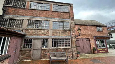 Image of a three story annexe made of red brick. Each floor has long, wooden sash windows, with a wooden barn door for the entrance of the building. Beside the annex a smaller red brick building has been built with a shop front on the bottom. Outside the annex is a wooden bench. 