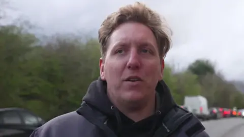 The image shows a man with light brown hair, wearing a dark hoodie standing beside a busy road 