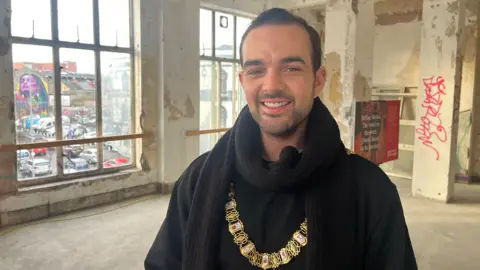 Belfast Lord Mayor Micky Murray - a man with short dark hair smiles at the camera. He is standing in the middle of an empty, concrete room with graffiti on the walls. He is wearing a black scarf, a black coat and top as well as the gold mayor chain.