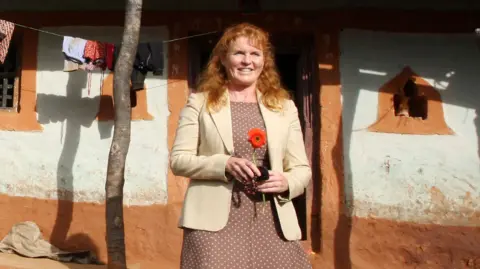 Getty Images Sarah Ferguson stands in front of a house with mud walls. She wears a beige jacket and brown dress, and carries a red flower. Some washing hangs on a line in the background.
