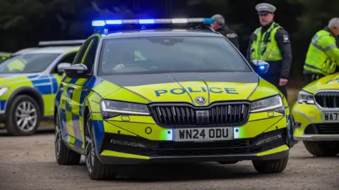 A black, yellow and blue police car drives off with its blue lights on from a car park while three officers stand by other police cars. Two officers are sat in the front of the car. It's number plate is WN24 0DU.