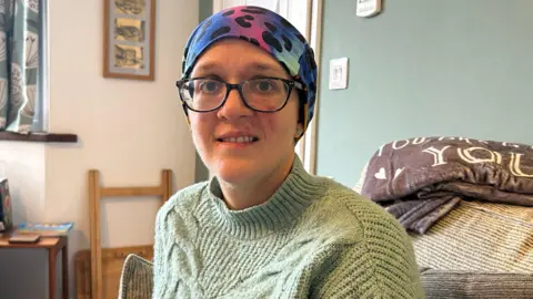 A woman wears a brightly-coloured headscarf and a green sweater. She is sitting on a sofa in a living room and has a red mark on her cheek.