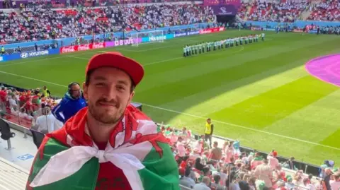 Rhodri, pictured in the crowd at a Wales football match wearing a Wales flag tied around his shoulders and red cap. The pitch is behind him. Rhodri has a moustache and stubble and is smiling at the camera. 