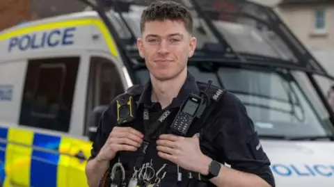 PC Thomas Fairbrother‑Wilcock has short dark brown hair and stands in full uniform in front a police van.