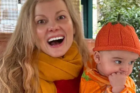 Lucy Turner Lucy is laughing and holding her baby in her arms. She has long blonde hair and he is wearing a pumpkin outfit including orange wool hat.