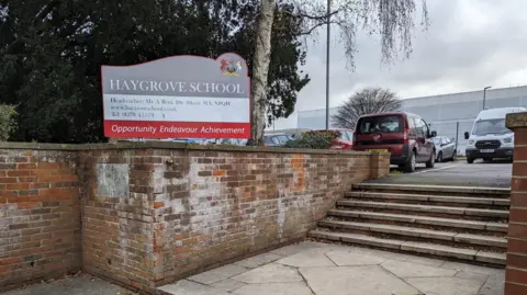 A sign outside Haygrove School bearing the name of the school on a grey background with a motto in red reading: "Opportunity, endeavour, achievement". There is a car park behind and the school building in the distance, which is covered up by a white material.
