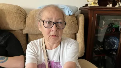 A woman in a white t shirt with a beige-strapped bag sitting on a tan sofa. A dark wood cabinet with a glass front can be seen to the right, containing a model car and a blue and white patterned bowl