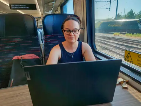 A woman with glasses wearing a navy blue vest sat on a train working on a laptop 