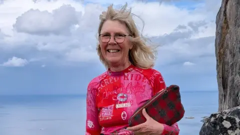 MND Association A woman smiling in front of the sea. She is wearing glasses and her blonde hair is blowing in the wind. She is also wearing a pink and red long sleeved cycling top, and is holding a red and green tartan clutch bag. The sky is cloudy and there is a sheer rock face on her left.