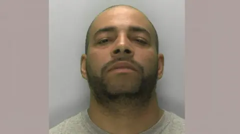 Gloucestershire Police Darren Atkins looking at the camera in a police mugshot. He has a light covering of stubble and a shaved head. He is wearing a grey sweatshirt