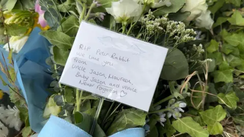A tribute written on white paper which reads RIP Peter Rabbit, we love you, from Uncle Jason, Maureen, Baby Jason and Venice. Never forgotten. The paper is in a bunch of flowers.