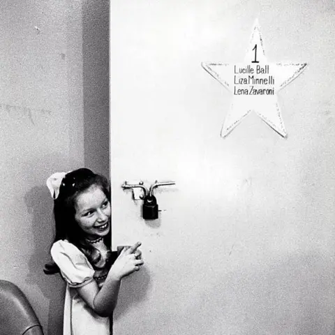 STAX/Specky Productions/BBC Scotland A black and white photo of Lena, in her trademark pinafore dress and bow, peeking out from behind her dressing room door. A star on the door lists its occupants - Lucille Ball, Liza Minelli and Lena Zavaroni.