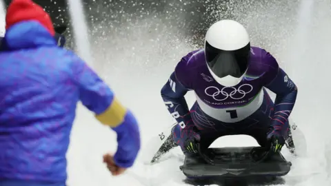 Reuters Matt Weston crosses the finishing line on his skeleton sled, wearing helmet and lycra Team GB kit. Another member of the team, in a red hat and blue jacket, appears at the left corner of the picture.