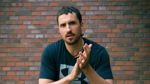 Pia Opp Andrew Moore wearing a black top rubbing his hands together against a brick wall background.