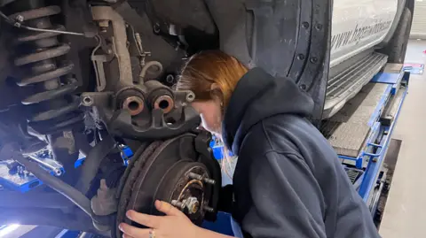 A close up of a female with ginger hair and a grey jumper who is replacing a car tyre.