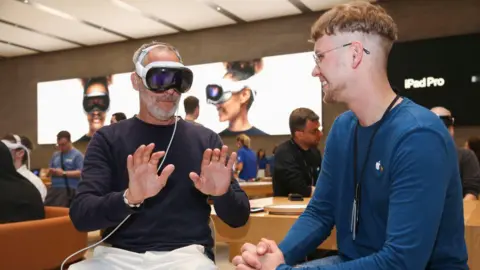 Getty Images A man sitting down wearing a white Apple Vision Pro headset on his head in an Apple store, which is connected with a white cable. He is holding his hands out in front of him. An Apple staff member is sitting next to him and smiling. 