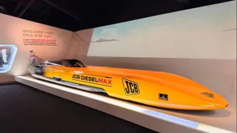 A long yellow JCB Dieselmax vehicle is displayed indoors in a museum setting, with information panels and a speed record graphic on the wall behind it.
