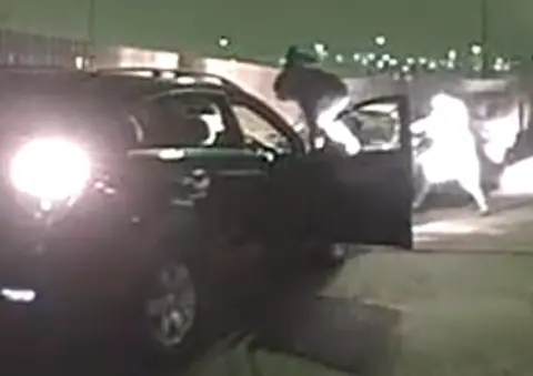 La policía de Cleveland muestra una imagen borrosa de las cámaras de seguridad en la que se ven varias personas corriendo alrededor de un coche negro con la puerta del conductor abierta.
