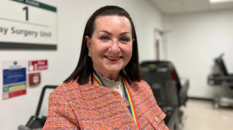 Deputy Barbara Ward in a hospital ward. She has black hair, and is wearing a pink and orange blazer with a rainbow coloured lanyard whilst smiling.
