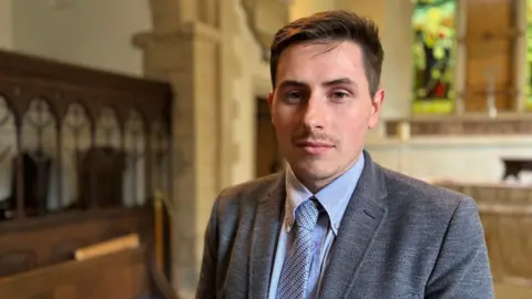 Samuel wearing a grey suit - blue shirt and a striped tie - standing in the church alter - looking at the camera.