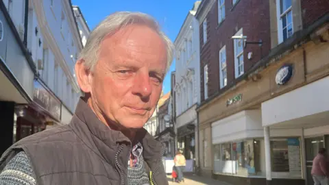 Ian Bucknall is wearing a grey bodywarmer over a patterned jumped. Behind him are city-centre shops, including Boots.