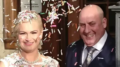 Anthony Webb and his wife Ewelina on their wedding day. The pair are both smiling and looking at the camera, with confetti surrounding their faces. He is bald and wears a blue suit, her blonde hair is tied back, and she wears a white peal necklace and wedding dress. 
