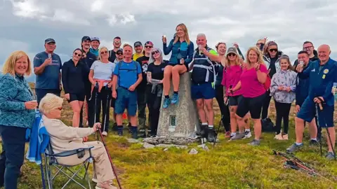 A group of around 20 people pose around a concrete trig point on a grassy hilltop under cloudy skies. Several people hold walking poles and drinks, while one person sits in a folding chair in the foreground. The landscape stretches into the distance behind the group, suggesting a hillwalking or summit gathering.