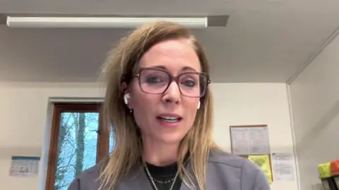 A woman with long blonde hair sits in a doctor's surgery. She is wearing glasses. She is talking into the camera and has earbuds in her ears.