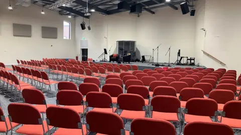 Chris Young/LDRS A large church hall with white washed walls and hundred of red chairs. At the front is a stage with set up for live music