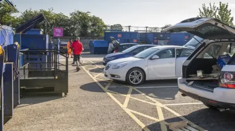 An outdoor area with several large metal blue containers. There are parked cars, some with their boots open displaying rubbish. 