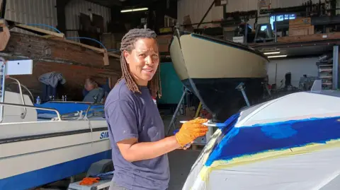 A woman stands in a boat yard building painting the hull of a small boat. She is smiling at the camera. She has short braids and is wearing a blue T-shirt. She is also wearing orange gloves. There are boats in the building behind her.