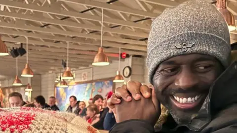 Alexis Brown Man in grey hat with black beard smiling off to the side with a busy bar or pub setting behind him.