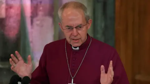 The Archbishop of Canterbury, dressed in a maroon cassock and a wearing a large silver cross on a chain, gestures with his hands as he speaks during the annual Lord Mayor's Banquet at Guildhall in London