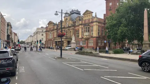A high street with old buildings on either side, some cars parked on the left, showing and people crossing the road further down. 