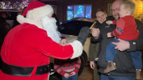 A couple hold their toddler while a man dressed as Santa talks to him