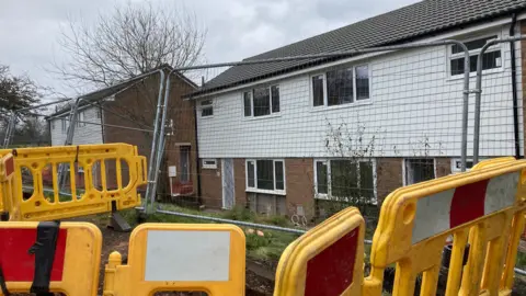 Yellow safety barriers and metal barriers are in the foreground with houses in the background which are being refurbished on Hospital Close in Leicester. 